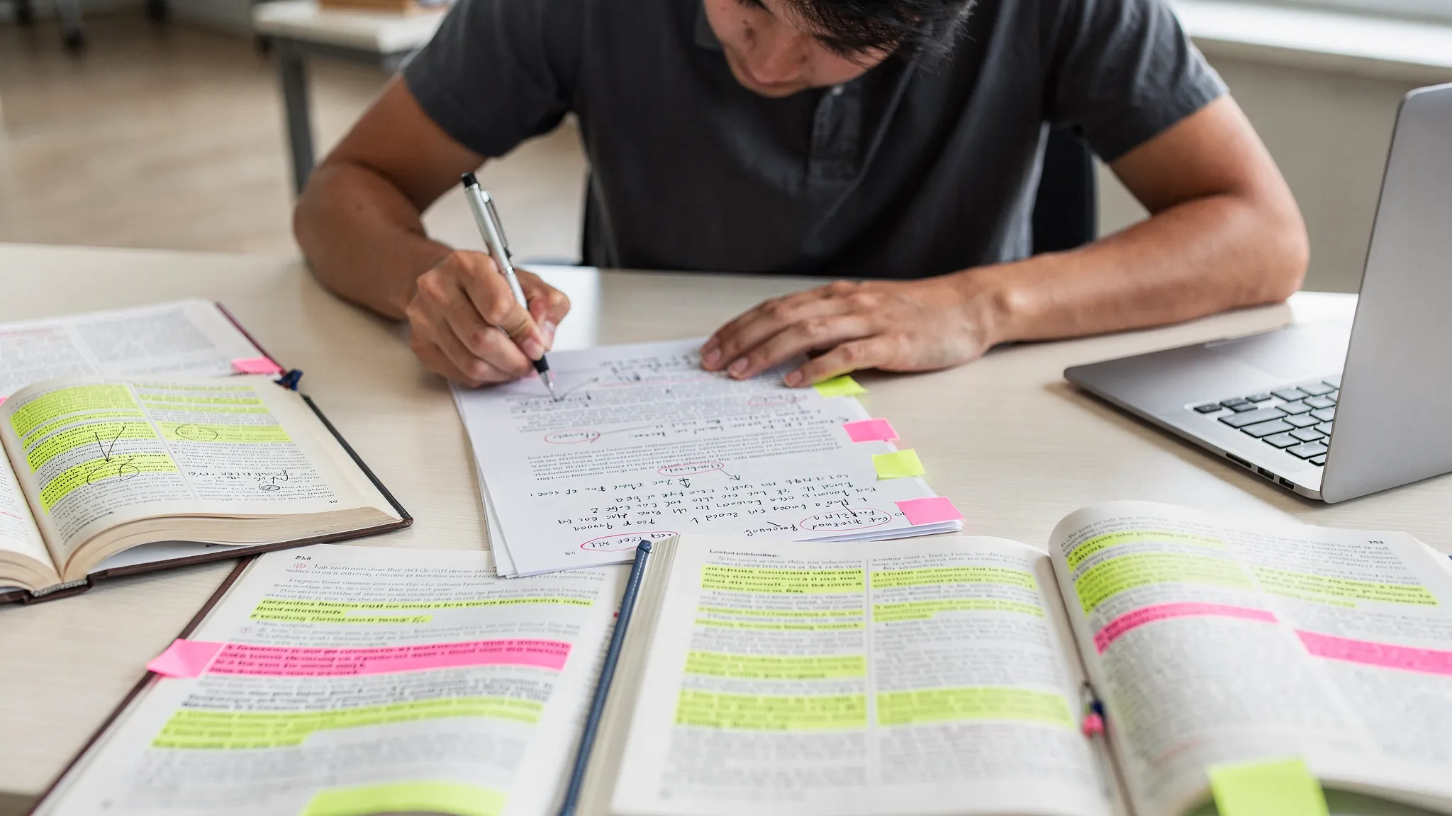 A student revising a printed draft with pen and sticky notes on a desk, with a laptop closed nearby and reference books and highlighted articles spread out, emphasizing an offline human editing process.