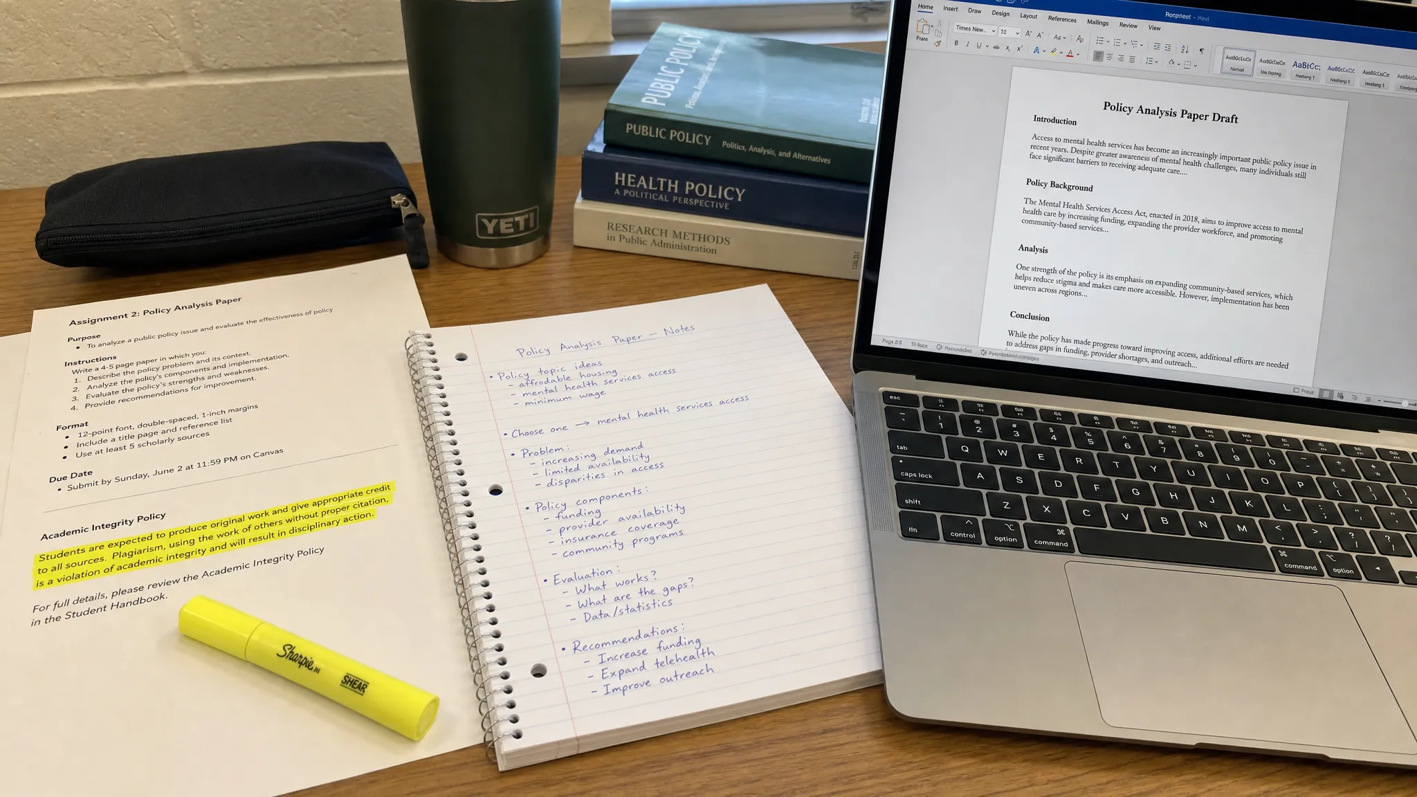A student desk with printed assignment instructions, handwritten notes, a highlighted academic integrity policy, and a laptop showing a draft document on the screen for review.