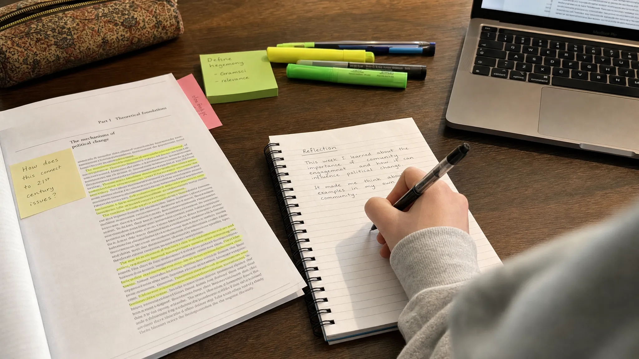 A person writing a short reflection in a notebook beside printed class readings, with highlighted passages, sticky notes, and a laptop pushed to the side, not as the main focus.