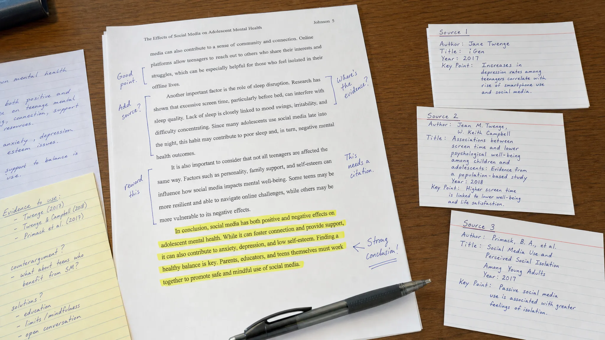 Printed essay pages on a desk with the conclusion paragraph highlighted in yellow, handwritten notes, source cards, and a pen beside the paper to show a careful review of writing evidence.