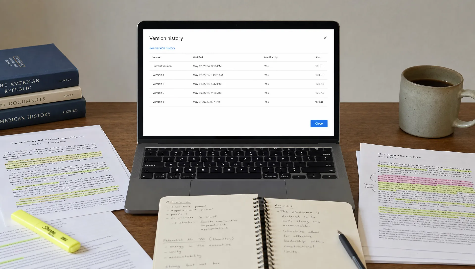 A study desk with printed essay drafts, handwritten notes, highlighted academic sources, and an open laptop facing the viewer that shows a simple document version history screen, with nothing displayed behind the laptop.